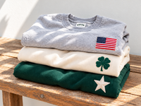 Stack of patch crewneck sweatshirts in heather grey, cream and hunter green with American flag, four leaf clover and star patches on a wooden bench