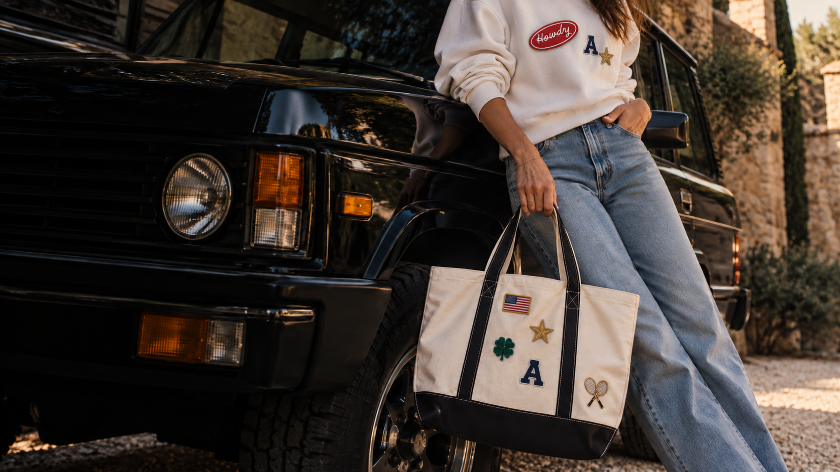 Woman in a cream Patch Crewneck with Howdy, letter and star patches, holding a navy Everyday Tote with flag, clover, star, letter and tennis patches