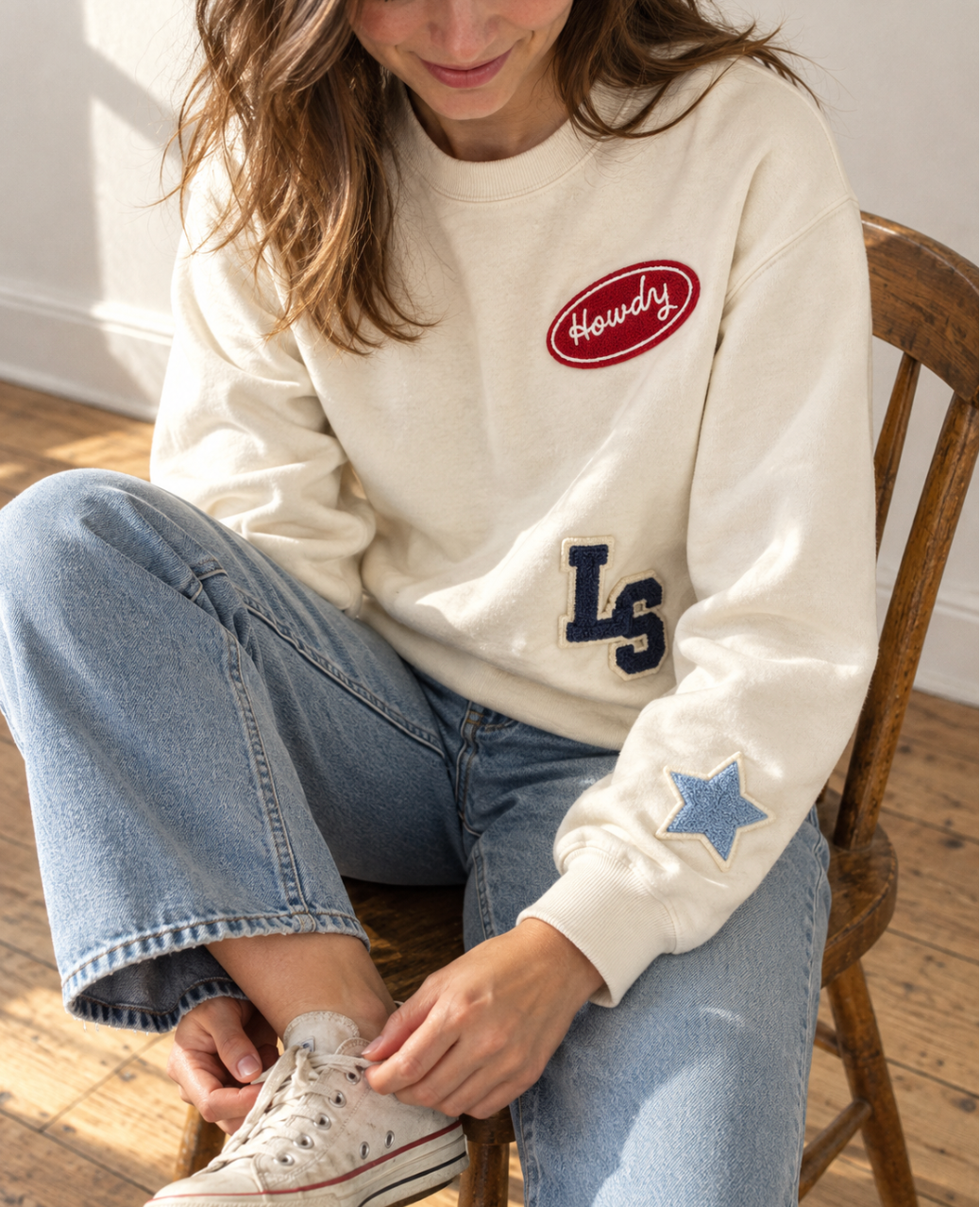 Woman sitting in a wooden chair wearing a cream patch crewneck sweatshirt with Howdy, LS monogram letters and a denim star patch
